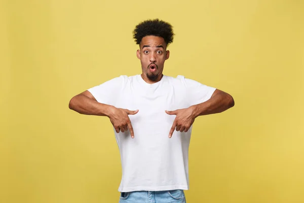 Astonished young African American man dressed in casual white shirt having excited fascinated look, pointing index finger at copy space on golden yellow background for your text or promotional content — Stock Photo, Image