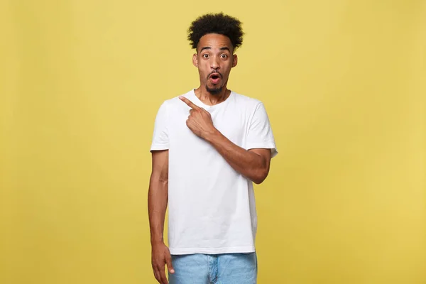 Astonished young African American man dressed in casual white shirt having excited fascinated look, pointing index finger at copy space on golden yellow background for your text or promotional content — Stock Photo, Image