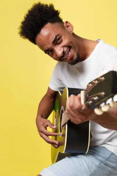 Young handsome African American retro styled guitarist playing acoustic guitar isolated on yellow gold background. — Stock Photo, Image