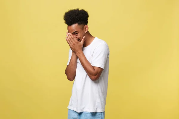 African american man with beard smiling having shy look peeking through fingers, covering face with hands looking confusedly broadly isolated over yellow background — Stock Photo, Image