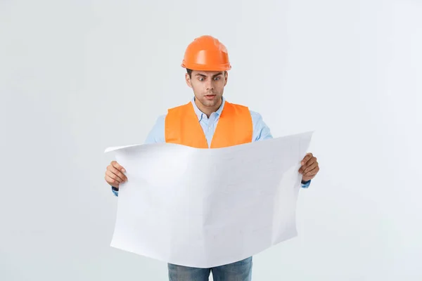 Εργαζόμενος κατασκευή σοκαρισμένος οικοδόμος κρατώντας στο χέρι ένα σχέδιο σχέδιο εγγράφου που απομονώνονται σε γκρίζο φόντο. — Φωτογραφία Αρχείου
