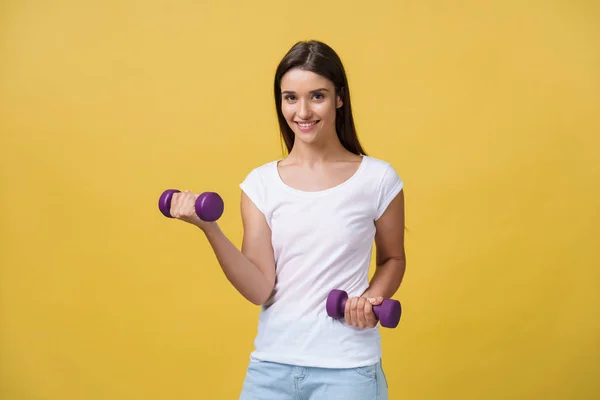 Shot of a beautiful and sporty young woman lifting up weights against yellow background. — Stock Photo, Image