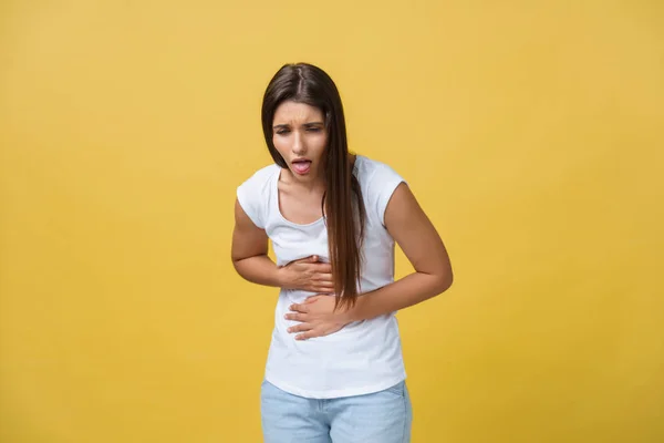 Retrato interior de una linda chica de pie con las manos cruzadas en el vientre, sintiéndose incómoda o sufriendo de dolor mientras mira a un lado, de pie sobre un fondo amarillo. Mujer tiene dolor de estómago —  Fotos de Stock