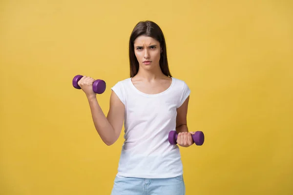 Feeling exhausted. Frustrated young woman in white shirt exercising with dumbbells and serious look while standing isolated on yellow background — Stock Photo, Image