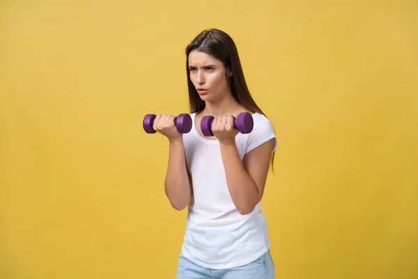 Feeling exhausted. Frustrated young woman in white shirt exercising with dumbbells and serious look while standing isolated on yellow background — Stock Photo, Image