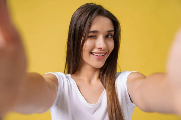 Agradable chica atractiva haciendo selfie en el estudio y riendo. Mujer joven guapa con cabello castaño tomando fotos de sí misma sobre un fondo amarillo brillante . — Foto de Stock