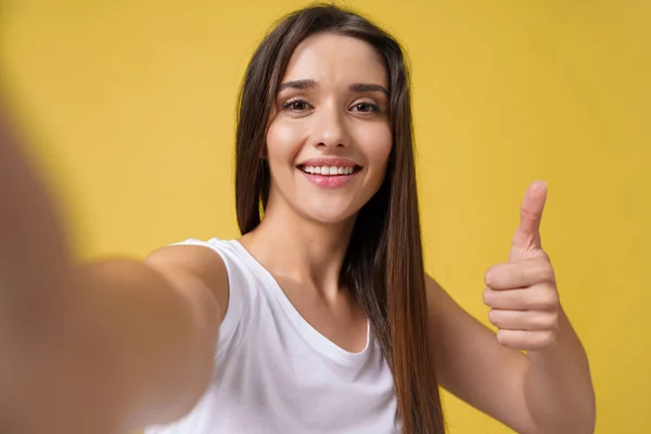 stock image Pleasant attractive girl making selfie in studio and laughing. Good-looking young woman with brown hair taking picture of herself on bright yellow background.