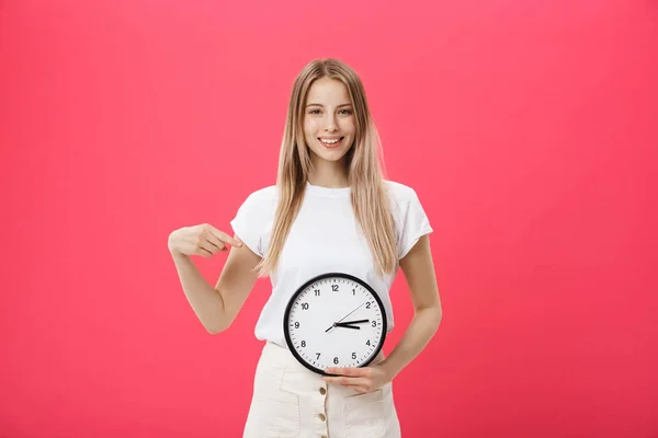 Portrait of an excited young girl dressed in white t-shirt pointing at alarm clock and looking at camera isolated over pink background