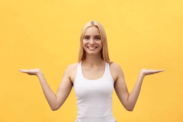Mujer joven hermosa y positiva emocional en una camisa blanca con una sorpresa en su cara mira hacia adelante, extendiendo sus manos hacia un lado contra una pared amarilla —  Fotos de Stock