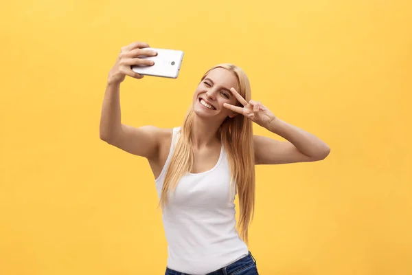 Αυτοπροσωπογραφία του πανέμορφο χαρούμενο κορίτσι γυρίσματα selfie στην μπροστινή κάμερα, χειρονομώ v-sign σύμβολο ειρήνης με τα δάχτυλα που απομονώνονται σε κίτρινο φόντο — Φωτογραφία Αρχείου