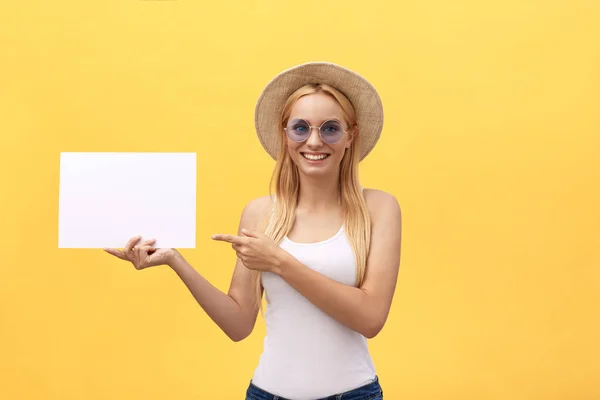 Mujer joven sobre fondo amarillo sosteniendo hoja de papel en blanco con la cara sorpresa apuntando espacio de copia del dedo . — Foto de Stock