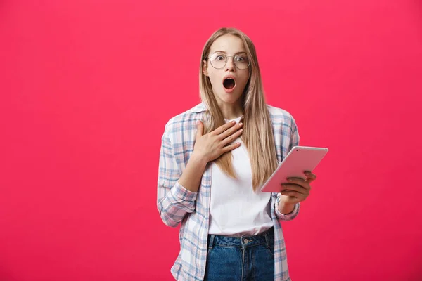Shocked woman looking at digital tablet with surprise and shock over pink background. Astonishment or hot news in internet