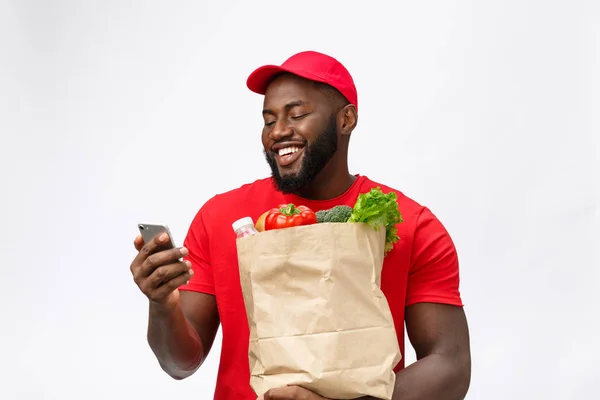 Dodací koncepce - Portrét Handsome Afroameričan doručovatel nebo kurýr s potravinářským balíčkem a mluvit na mobilním telefonu zkontrolovat objednávku. Izolováno na pozadí studia Grey — Stock fotografie