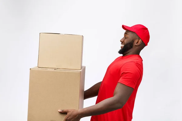 Concept de livraison - Vue de côté Portrait du livreur afro-américain heureux en tissu rouge tenant un paquet de boîte. Isolé sur fond gris. Espace de copie — Photo