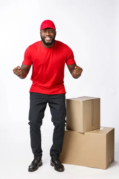 Concept de livraison - Portrait du livreur afro-américain heureux en tissu rouge tenant un paquet de boîte. Isolé sur fond de studio Grey. Espace de copie — Photo