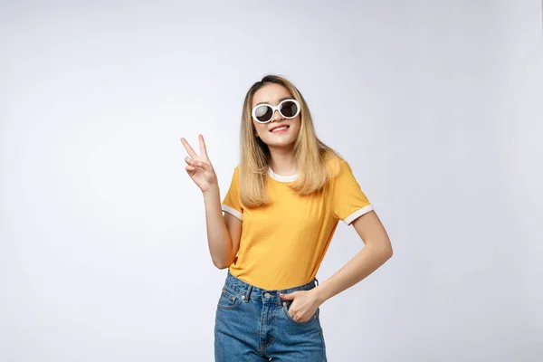 Genç Asyalı kadın üzerinde güneş gözlüğü takıyor gösterilen arka plan izole ve parmakları ile işaret kendinden emin ve mutlu gülümseyerek ise ikinci. — Stok fotoğraf