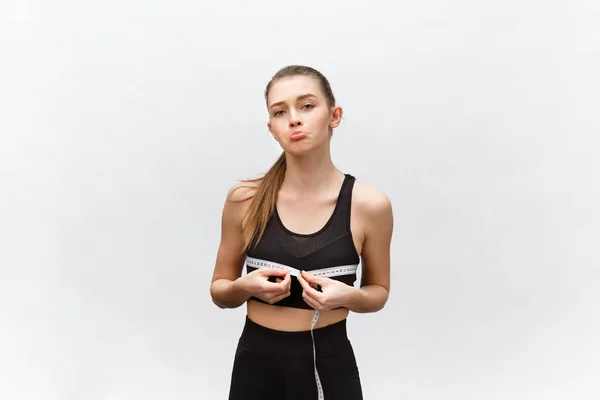 Young woman with excess weight in sporty top sadly looking on result of measuring waist over white background — Stock Photo, Image