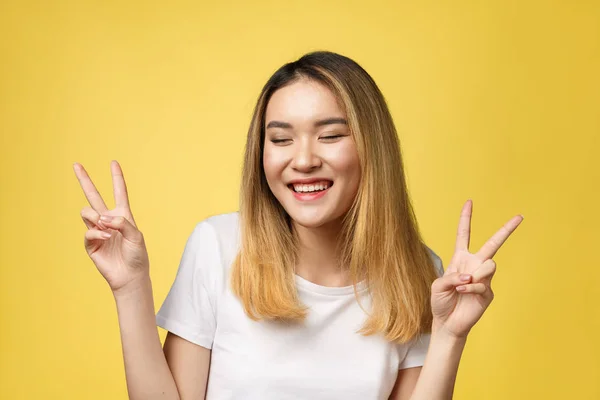 Agradable mujer asiática en camiseta mostrando gestos de paz y mirando a la cámara sobre fondo amarillo —  Fotos de Stock