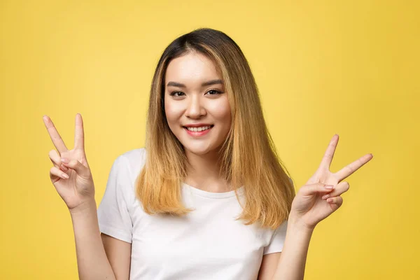 Agradável mulher asiática em t-shirt mostrando gestos de paz e olhando para a câmera sobre fundo amarelo — Fotografia de Stock