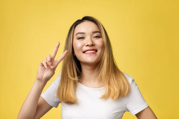 Piacevole asiatica donna in t-shirt mostrando gesti di pace e guardando la fotocamera su sfondo giallo — Foto Stock