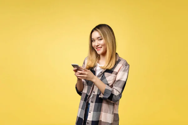 Imagen de mujer asiática satisfecha con el pelo largo sonriendo y mensajes de texto en el teléfono celular sosteniendo en la mano aislado sobre fondo amarillo — Foto de Stock