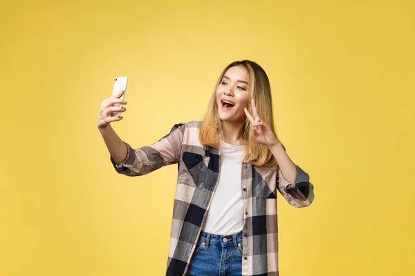 Pretty girl take a self portrait with her smart phone. Asian girl selfie, isolated on yellow background. — Stock Photo, Image