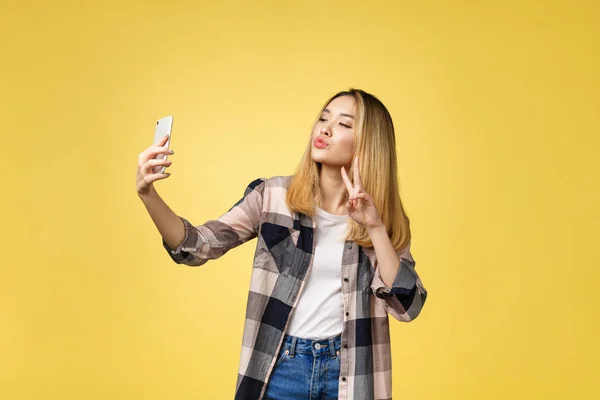 Söt flicka ta ett självporträtt med sin smarta telefon. Asiatisk flicka selfie, isolerad på gul bakgrund. — Stockfoto