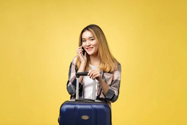 Ritratto di felice viaggiatore femminile asiatico con valigia e guardando il cellulare sullo sfondo giallo isolato — Foto Stock