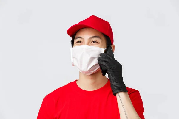 Covid-19, concept d'achat et de livraison en ligne auto-quarantaine. Amical jeune asiatique courier en masque médical et gants de protection, appelant le client, répondre au téléphone du client, souriant, fond gris — Photo