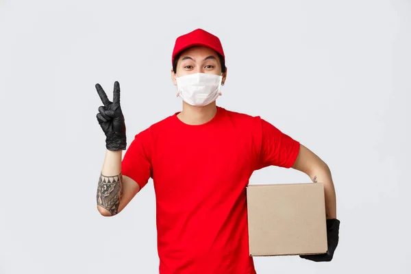 Έννοια της παράδοσης και φορείς κατά τη διάρκεια της πανδημίας coronavirus. Cute courier in red cap, t-shirt and medical mask with gloves, show peace sign holding box to delivery client, αποστολή online παραγγελία σας — Φωτογραφία Αρχείου