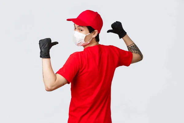 Visão traseira do asiático entregador em máscara médica e luvas de proteção, use boné vermelho, t-shirt, vire-se para a câmera, apontando para trás para mostrar o logotipo da empresa em uniforme, logística, correio e conceito de compras — Fotografia de Stock