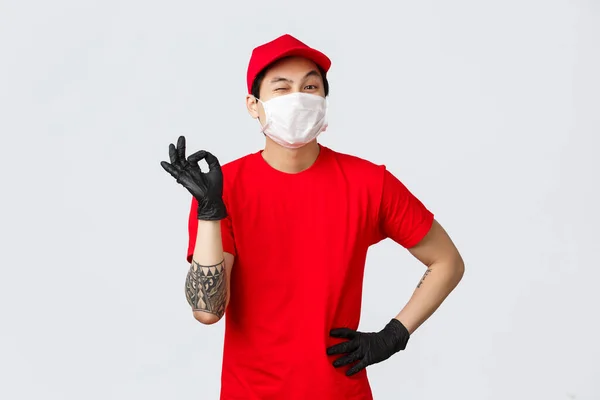 Pas de problème, livrez votre colis. Joyeux livreur asiatique en uniforme rouge, montrer signe correct et clin d'oeil, assurer la sécurité du colis. Portez des gants de protection et un masque médical, restez en sécurité et magasinez en ligne — Photo