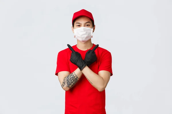 Mladý sebevědomý doručovatel v červeném tričku a čepici, nosit ochranné rukavice a lékařskou masku doručit balíčky domů zákazníka během covid-19, ukazuje varianty, ukazuje stranou, dvě možnosti — Stock fotografie