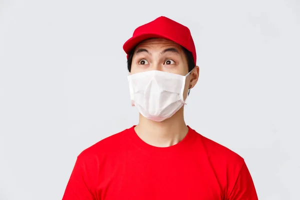 Ковид-19, самокарантинный онлайн-шопинг и концепция доставки. Возбужденный и впечатленный курьер в медицинской маске и униформе красной шапки, футболке, глядя в левый верхний угол изумленный, серый фон — стоковое фото