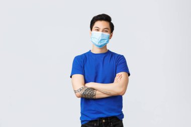 Farklı duygular, sosyal mesafeler, koronavirüs karantinası ve yaşam tarzı konsepti. Mavi tişörtlü, tıbbi maskeli, göğüs göğsünde çapraz göğüslü yakışıklı Asyalı çocuk, evde pozitif kal.