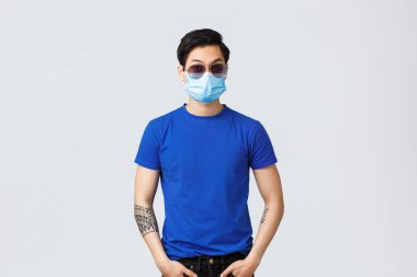 Covid019 yaşam tarzı, insan duyguları ve karantina kavramı üzerine eğlence. Güneş gözlüklü, tıbbi maskeli, maço ya da güvenlik görevlisi gibi davranan küstah ve havalı Asyalı adam gri arka planda duruyor.