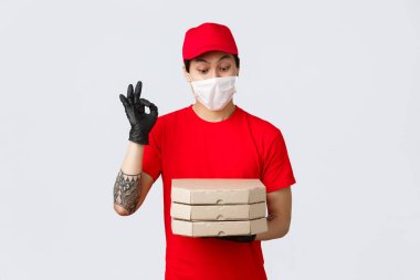 Eğlendirilmiş Asyalı kurye kırmızı üniformalı, kep ve tişörtlü, müşteri güvenliği için koruyucu eldiven ve tıbbi maske takıyor, kapıya yemek getiriyor, pizza getiriyor, tamam işareti yapıyor ve lezzetli pizzaya bakıyor.