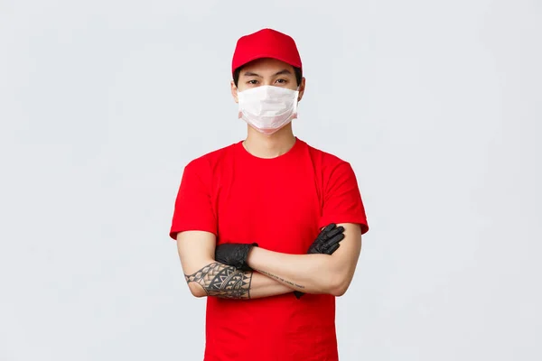 Magabiztos, fiatal ázsiai férfi, piros kézbesítési egyenruhában és sapkában, orvosi maszkot és kesztyűt visel, készen áll az áru átadására. Biztonságos vásárlási koncepció. Futárkarok a mellkason — Stock Fotó