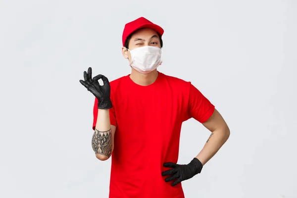 Žádný problém, doručte zásilku. Veselý asijský poslíček v červené uniformě, ukázat dobré znamení a mrknout, zajistit bezpečnost balíčku. Noste ochranné rukavice a lékařskou masku, buďte v bezpečí a nakupujte online — Stock fotografie