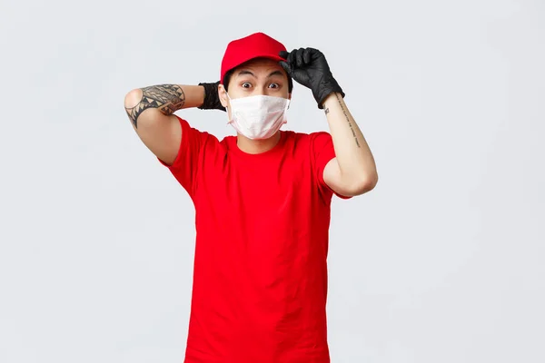 Nerwowy azjatycki dostawca pospiesznie dostarcza towar, zakłada mundur w czerwonej czapce, maskę medyczną i rękawiczki ochronne, wysyła paczki podczas pandemii Kowid-19. Pojęcie zakupów online i samoizolacji — Zdjęcie stockowe