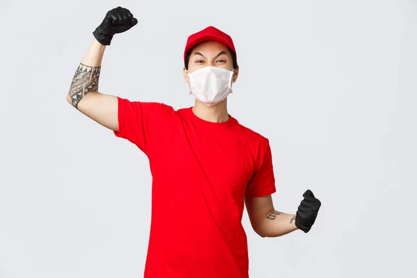 Wesoły entuzjastyczny dostawca w czerwonej koszulce i czapce, ubrany w maskę medyczną, rękawiczki ochronne, podnosząc rękę do góry śpiew, powiedzieć tak lub hura, świętować sukces, zachęcić do porządku online — Zdjęcie stockowe
