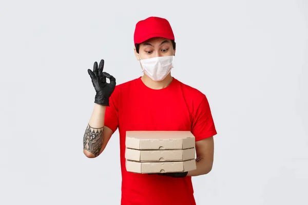 Zábavná asijská dodávka muž červená uniforma, čepice a tričko, nošení ochranných rukavic a lékařské masky pro bezpečnost klientů, jak dodat jídlo ke dveřím, přinést pizzu, ukázat v pořádku znamení a zírat na chutnou pizzu — Stock fotografie