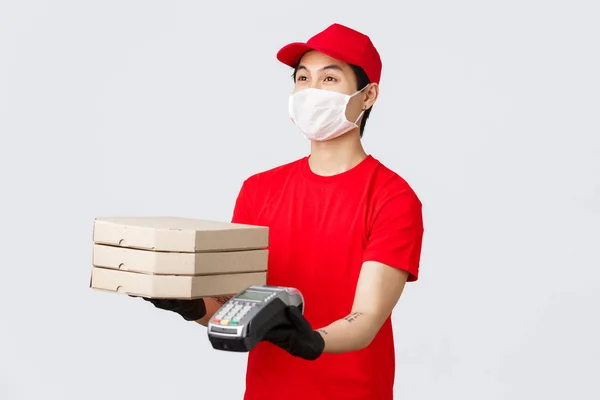 Consegna senza contatto, acquisto sicuro e shopping durante il concetto di coronavirus. Gentile corriere in cappuccio uniforme rossa e t-shirt, dando ordine di consegna pizza cliente e terminale di pagamento, sfondo grigio — Foto Stock