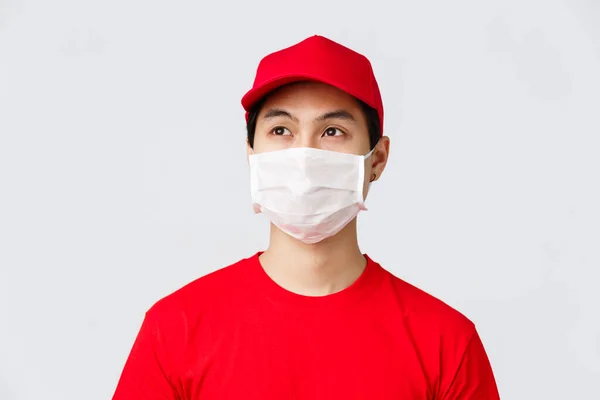 Covid-19, self-quarantine online shopping and delivery concept. Thoughtful young asian courier working during coronavirus outbreak to bring orders client doorstem, wear medical mask and uniform — Stock Photo, Image