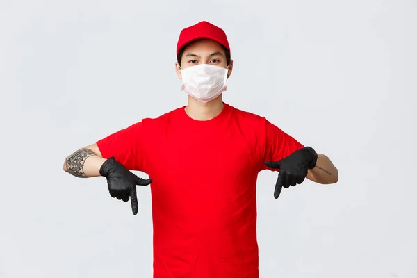 Look here. Serious, confident asian delivery guy with tattooed arm, pointing fingers down to show advertisement promo in protective gloves and medical mask. Courier wearing uniform, delivery parcels — Stock Photo, Image