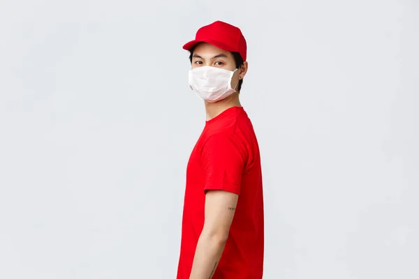 Portré ázsiai futárról piros sapkában és pólóban, védőorvosi maszkot visel, és szemtől szemben kamerázik. Az egyenruhás futár csomagokat szállít, míg az emberek biztonságban maradnak otthon a 19 karantén alatt. — Stock Fotó