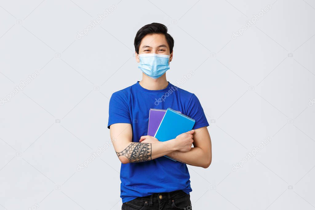 Back to school, studying during covid-19, education and university life concept. Happy young male student in medical mask, holding notebooks, homework and smiling, stand grey background