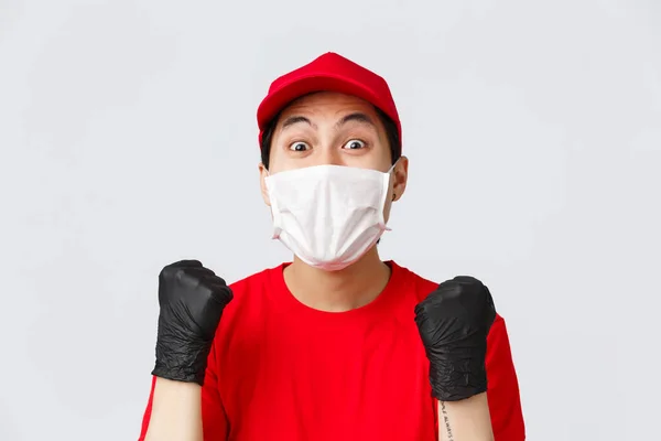 Covid-19, self-quarantine online shopping concept. Hopeful and relieved delivery guy in red cap, medical mask and gloves, fist pump achieve goal, stare amazed, received raise in courier service
