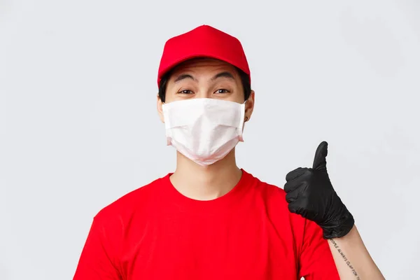 Covid-19, self-quarantine online shopping and delivery concept. Cheerful asian courier in medical mask and protective gloves, wear red uniform, show thumb-up in approval, recommend make safe orders — Stock Photo, Image