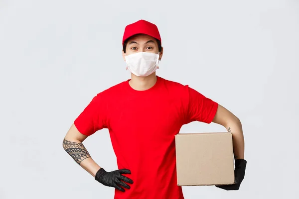 Vidám ázsiai férfi piros sapkában és pólóban, csomagtartó dobozban, védőkesztyűben és orvosi maszkban. Futárfelvevő csomag az ügyféltől, szállítási doboz a covid-19 járvány kitörésekor — Stock Fotó